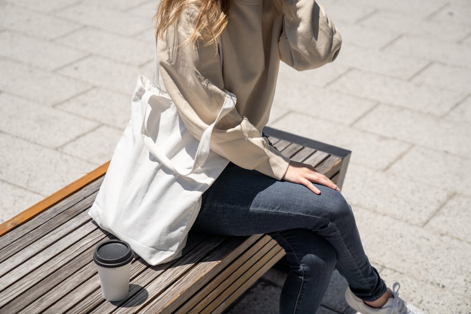 Person sitting on a bench with coffee wearing denim jeans and holding a tote bag.