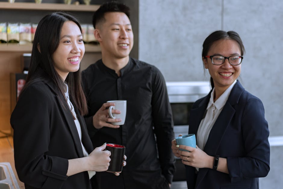 Three Asian coworkers enjoying a relaxed coffee break indoors, smiling and holding mugs.