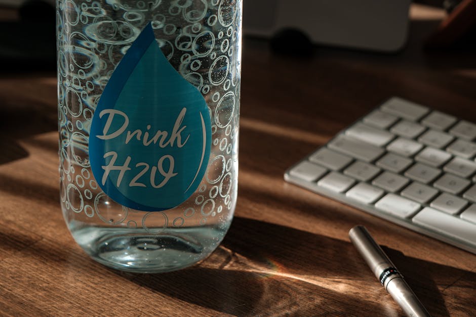 Glass water bottle labeled 'Drink H2O' on an office desk with sunlight and keyboard.