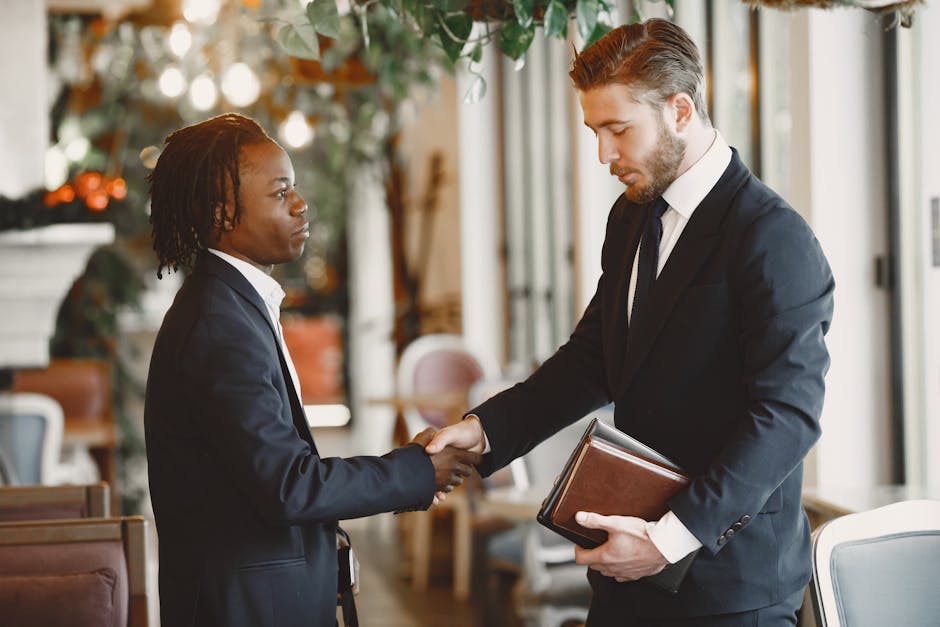 Two businessmen shake hands in a professional setting. Indoors, formal attire.